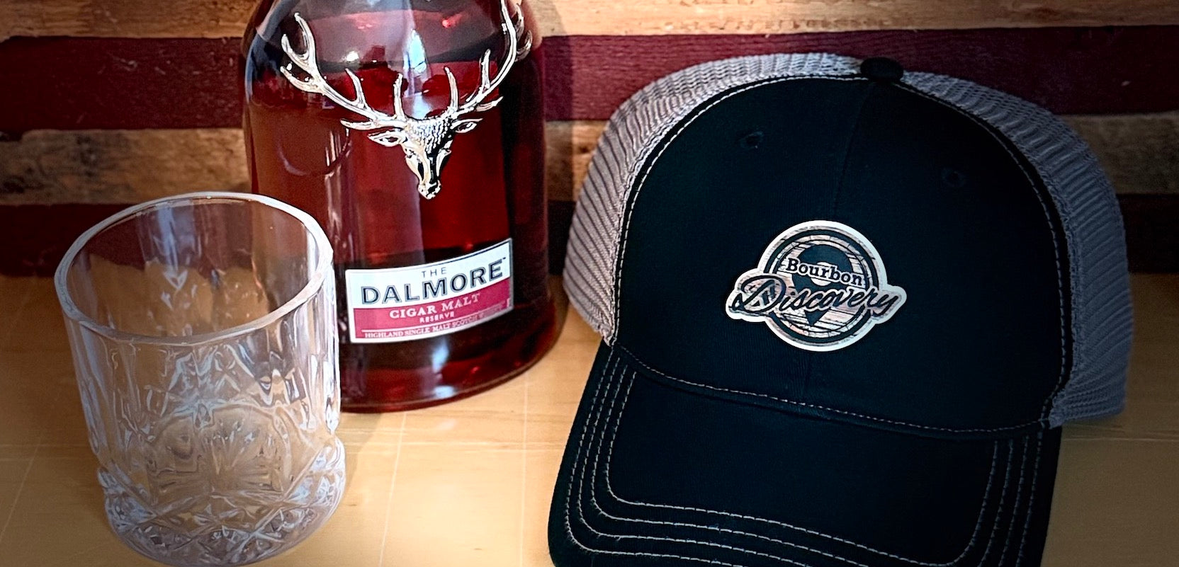 Whisky bottle, glass, and black cap with custom logo patch on a wooden surface
