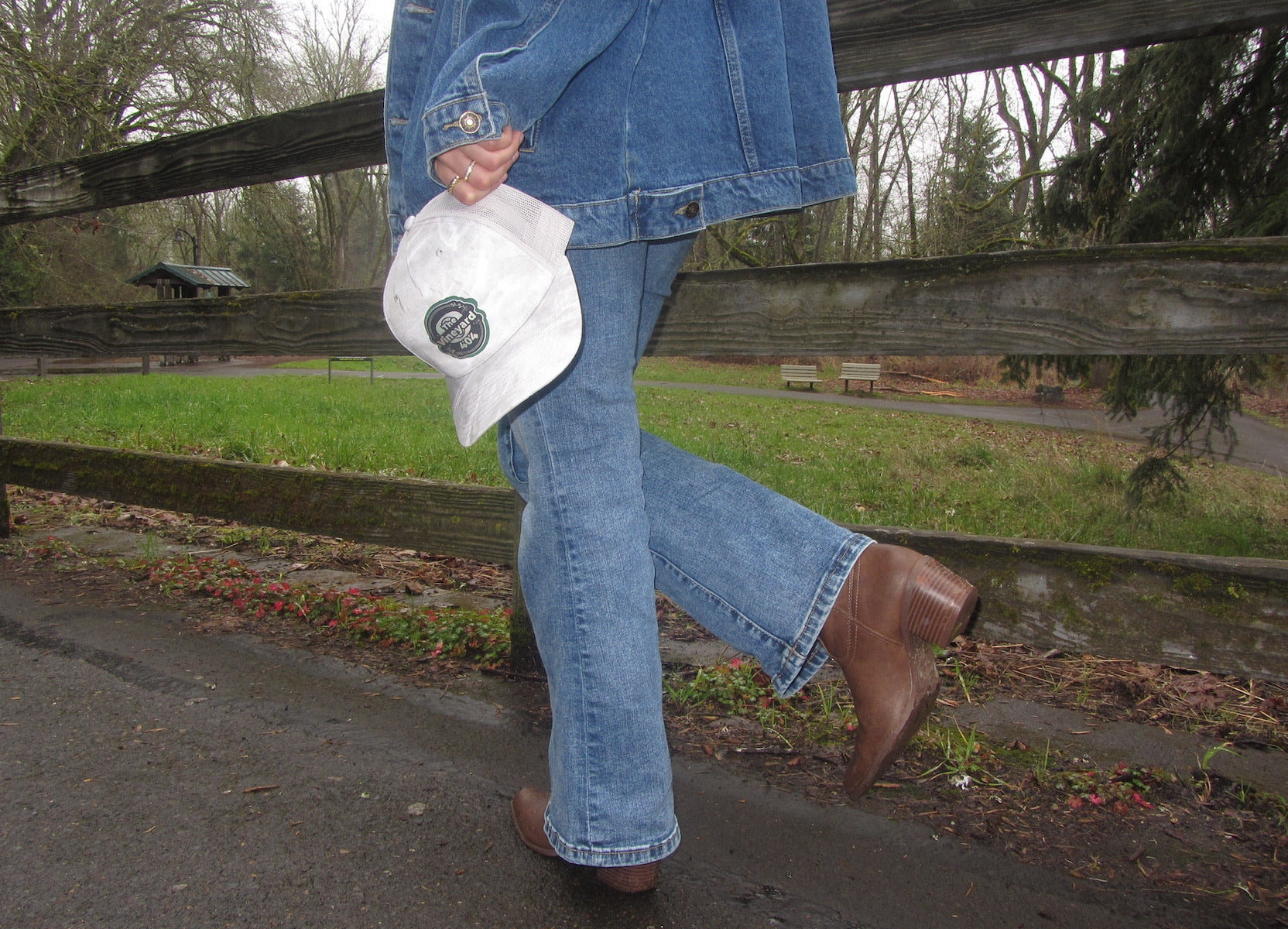 Woman holding a white cap with a custom logo, standing near a wooden fence in an outdoor setting