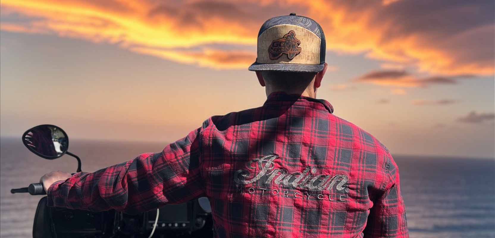 Man wearing a custom baseball cap, sitting on a motorcycle with a sunset over water in the background