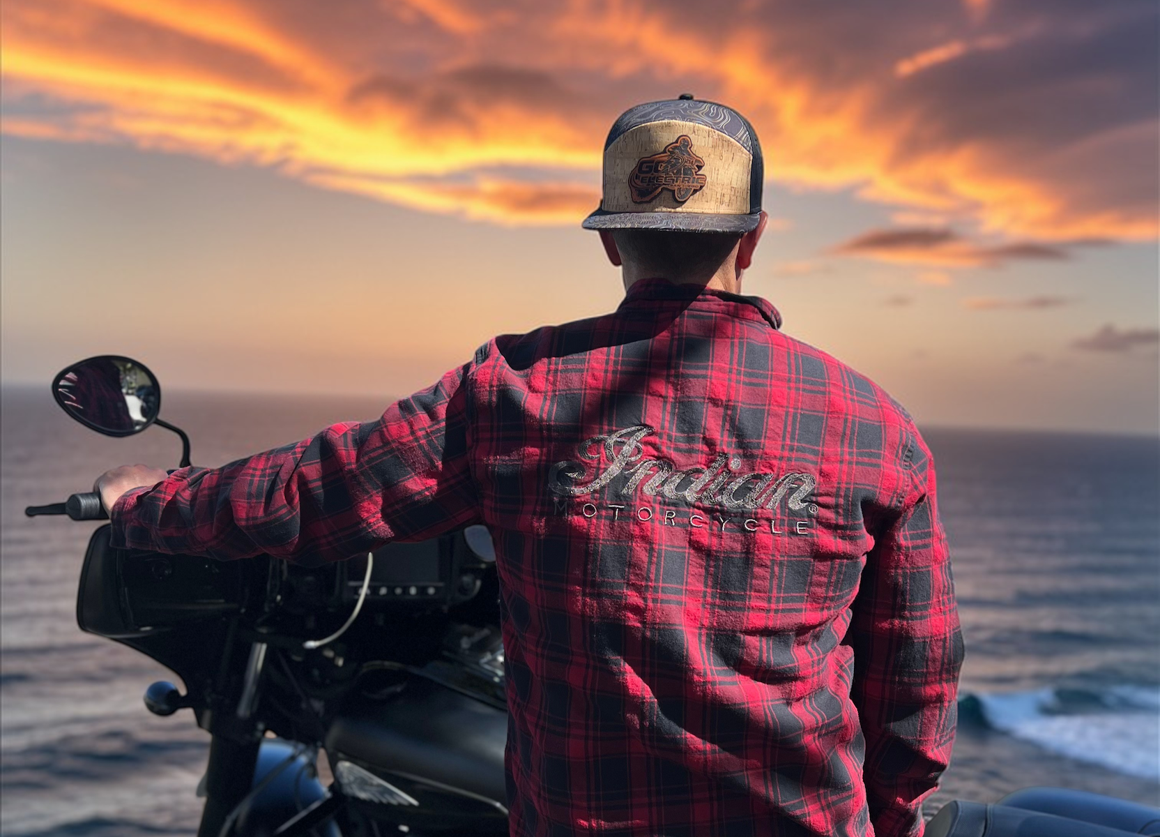 Man wearing a custom baseball cap, sitting on a motorcycle with a sunset over water in the background