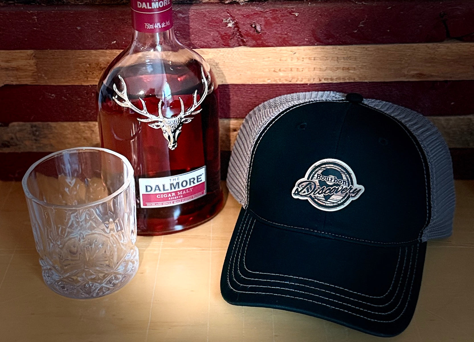 Whisky bottle, glass, and black cap with custom logo patch on a wooden surface